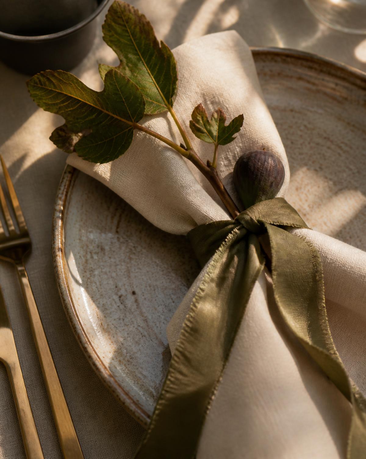 Place setting with ivory linen napkin tied in olive silk ribbon, fig branch and brass flatware